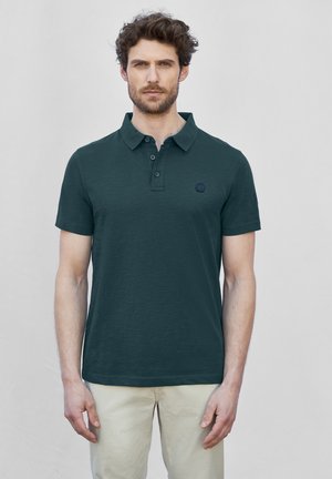 Dunkelgrünes Poloshirt aus strukturiertem Stoff, mit Kragen, drei Knöpfen und einem kleinen Logo auf der linken Brust. Kurzarm.