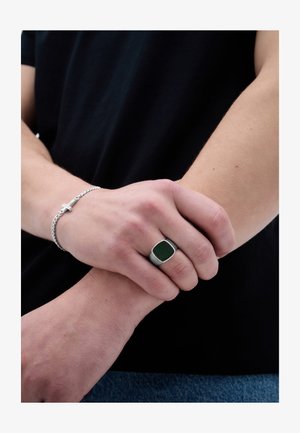 Henkilö, joka pitää oikealla kädellään vasenta rystystään esillä hopeinen ristikoru ja mustalla kellotaululla varustettu hopeasormus.