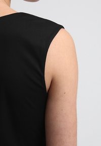 Černý bezrukávový top s hladkou texturou, který má kulatý výstřih a minimalistický design. Detailní záběr na rameno a horní část zad.