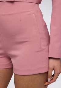 Shorts rosa su misura realizzati in tessuto liscio, con tasche laterali e un taglio pulito. La texture appare morbida con un design strutturato.