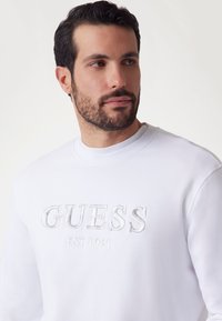 Biała bluza z haftowanym napisem "GUESS" i szczegółem "EST. 1981". Gładki materiał, okrągły dekolt i długie rękawy.