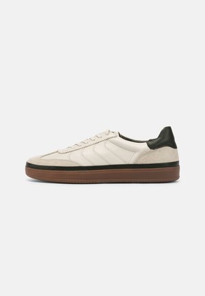 Lace-up sneaker in cream leather and beige suede, featuring a green heel accent, textured stripes, and a brown rubber sole.