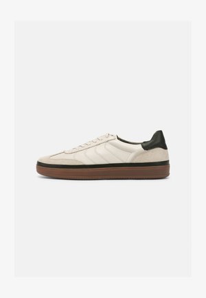 Basket en cuir crème et suède beige, avec un accent de talon vert, des rayures texturées et une semelle en caoutchouc brun.