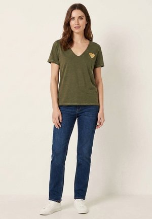 Femme portant un t-shirt vert olive à col en V avec un cœur doré sur la poitrine gauche, jean bleu et baskets blanches, debout devant un fond uni.