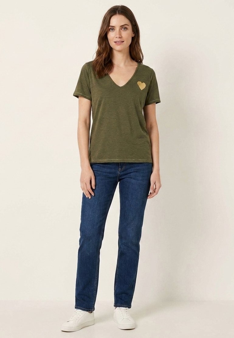 Femme portant un t-shirt vert olive à col en V avec un cœur doré sur la poitrine gauche, jean bleu et baskets blanches, debout devant un fond uni.