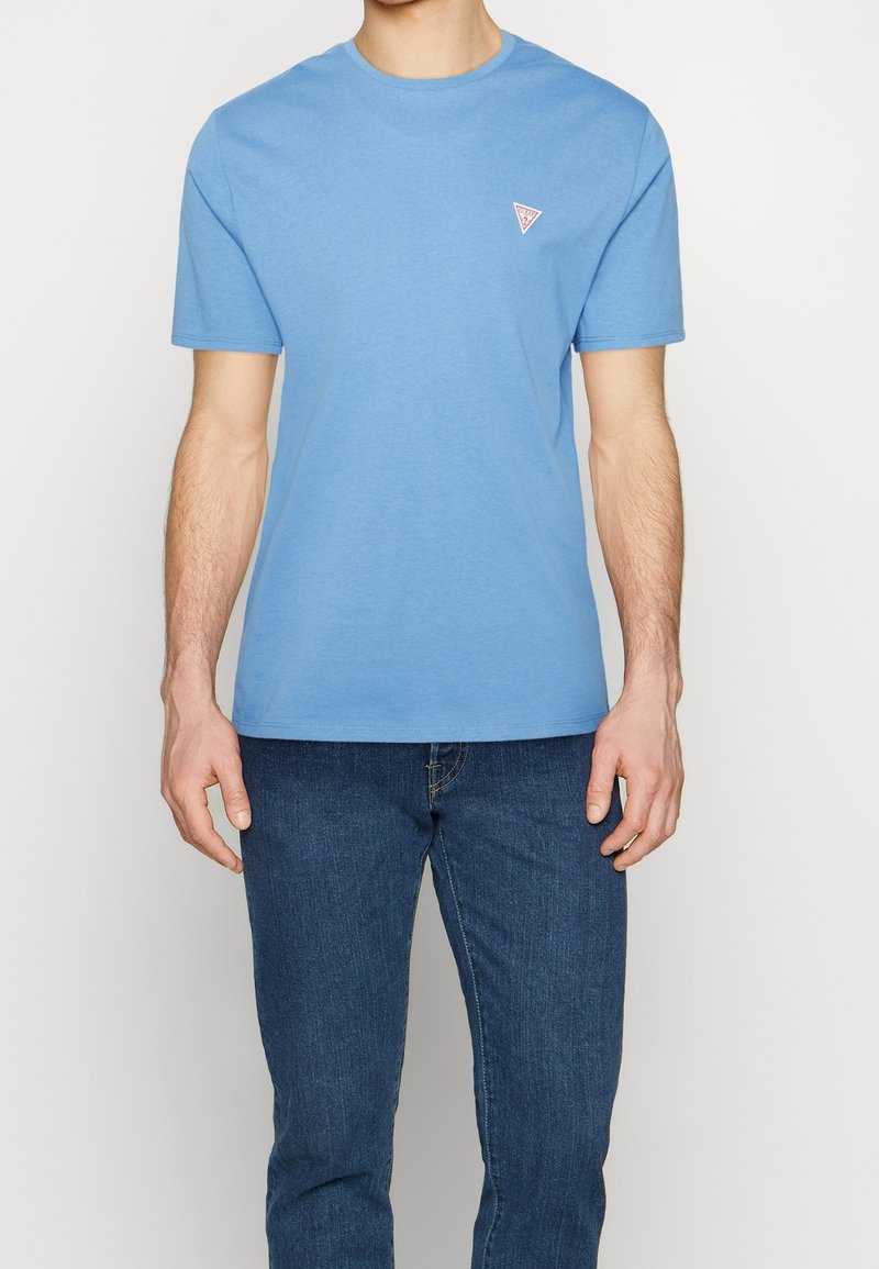T-shirt en coton bleu clair avec un col rond et des manches courtes, arborant un petit logo triangulaire sur le côté gauche de la poitrine.