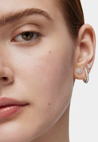 Primer plano de una oreja izquierda con tres pendientes plateados: un pendiente con forma de oso de peluche, un pequeño pendiente también con forma de oso de peluche y un aro curvado.