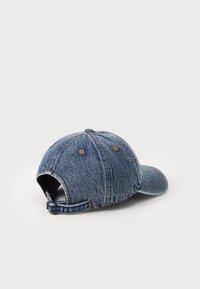 Denimkasket med en struktureret form, blå farve og en let afdæmpet tekstur. Har et justerbart spænde bagpå og kontraststing.