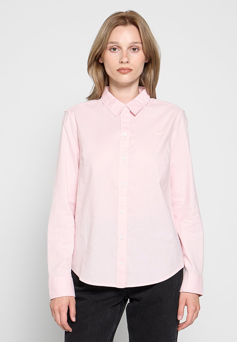 Camisa de color rosa claro con cuello y mangas largas, que presenta un cierre de botones en la parte delantera y un dobladillo curvado. Elaborada con una tela ligera y de textura sutil.