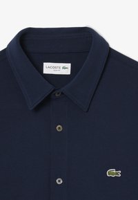 Mørkeblå slim-fit polo skjorte laget av strukturert stoff. Har krage, to knapper og en liten grønn krokodillelogo på brystet.