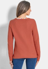 Frau mit langen grauen Haaren, die einen strukturierten rostoranfarbenen Pullover und blaue Jeans trägt, mit dem Rücken zu einer schlichten hellen Hintergrund.