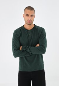Tume rohelise värvi pikkade varrukatega henley särk, mille kaeluses on nööpdetailid ja keha lähedal istuvad varrukad. Pehme tekstuur ja vabam lõige.