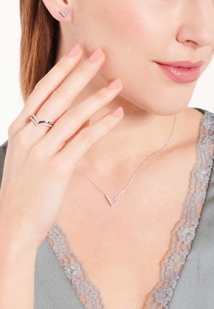 Silver V-shaped necklace and matching earrings, set against a soft gray top, with a subtle lace detail. Smooth texture and clean design.