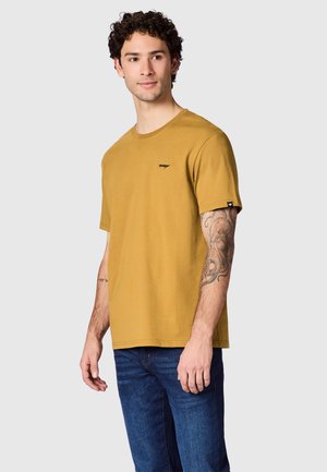 Mężczyzna z kręconymi włosami, w musztardowo-żółtej koszuli z krótkim rękawem i niebieskich dżinsach, pokazuje wytatuowane prawe przedramię, patrzy lekko w lewo.