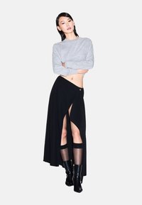 Vrouw die met gekruiste armen staat, gekleed in een lichtgrijze sweater, een zwart asymmetrische rok met een hoge split, transparante kniekousen en zwarte enkellaarsjes.