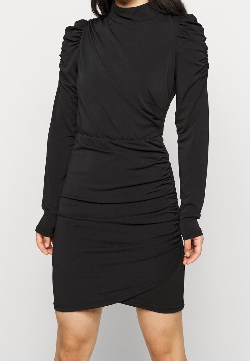 Femme portant une robe noire à manches longues avec des épaules et une taille froncées, un col montant et une jupe portefeuille.