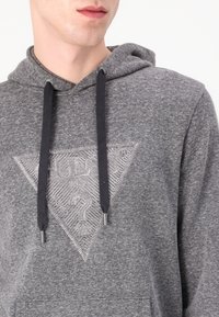 Graue Kapuzenpullover mit einem dreieckigen Grafikdesign, das ein strukturiertes Muster und ein gesticktes Logo zeigt. Schwarze Kordeln mit Metallenden; weiches, lässiges Material.