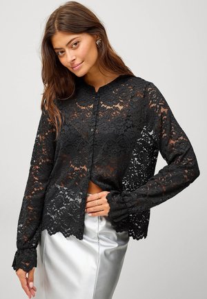 Zwarte kanten blouse met lange mouwen, ronde kraag, bloempatroon, knoopsluiting, en uitlopende manchetten. Gecombineerd met een zilveren rok.