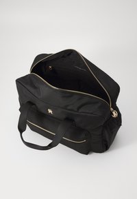 Sac fourre-tout en tissu noir avec fermeture éclair dorée et logo. Il présente deux poignées supérieures et un intérieur ouvert. Texture lisse, forme rectangulaire.