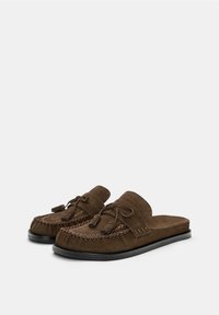Mocassins en suède brun à enfiler avec des gland, des détails de couture tressée et des semelles noires, inclinés sur un fond blanc.