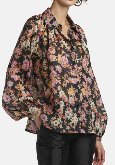 Donna che indossa una camicetta nera trasparente con stampa floreale rosa, gialla e bianca, maniche a sbuffo e bottoni neri, abbinata a pantaloni neri.