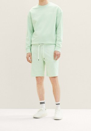 Osoba ubrana w jasnozieloną bluzę z kapturem i pasujące krótkie spodenki z białym sznurkiem, białe trampki oraz białe skarpety z czarnymi paskami, stojąca.