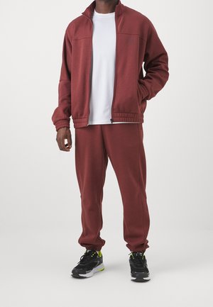 Man wearing maroon zip-up jacket and matching sweatpants with black and neon green sneakers, standing with one hand in pocket.