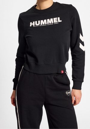 Personne portant un sweat-shirt et un pantalon de sport Hummel noirs avec des logos blancs et des chevrons sur la manche et le côté du pantalon.