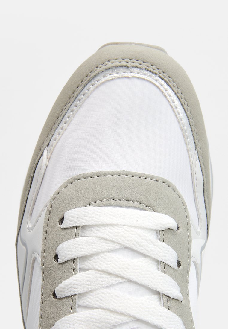 Zapatilla blanca y gris con una parte superior de tela suave y acentos de ante, que presenta cordones planos blancos y un diseño de punta redondeada.