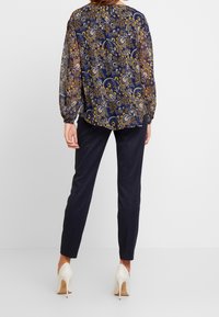 Blusa fluida navy con motivo paisley dorato, maniche lunghe e polsini elastici; abbinata a pantaloni neri attillati e tacchi alti bianchi.