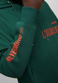 Sweat à capuche vert en mélange de coton, avec le texte orange "NEVERMIND 1991" sur la manche et la poitrine, doté de poignets côtelés et d'une poche frontale kangourou.