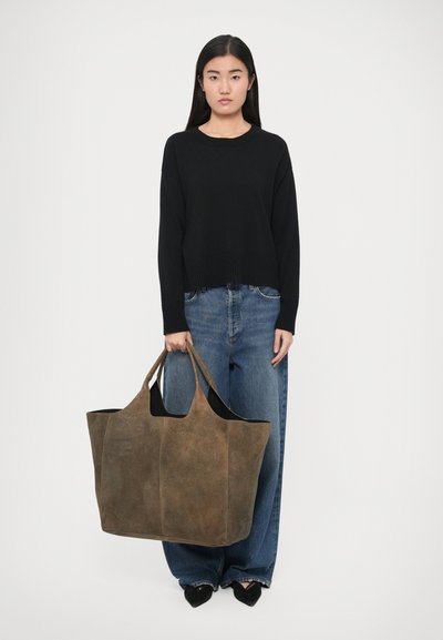 Geantă tote maro din piele nubuc, cu deschidere largă, două mânere și formă structurată. Model purtând un pulover negru și blugi denim largi.