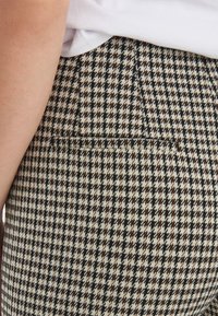 Kostkované kalhoty se vzorem houndstooth v černé, krémové a hnědé barvě, se zadní kapsou a viditelnou texturou látky poblíž pasu.