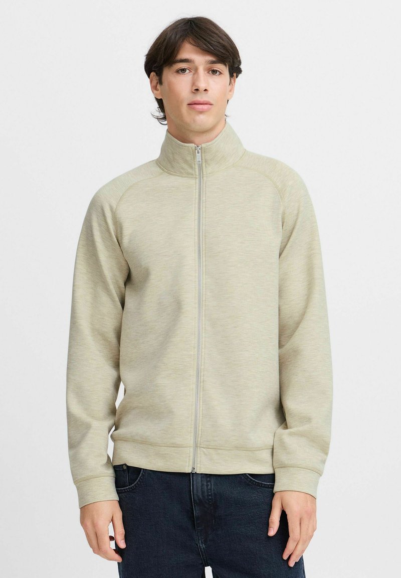 Light beige zip-up sweatshirt with a high collar, ribbed cuffs, and structured seams, worn over dark jeans. Soft fabric with a textured appearance.