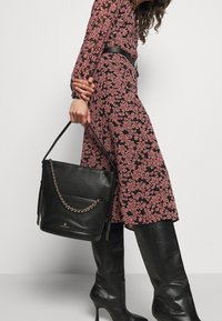 Musta tekstiilipintainen nahkakassi, jossa on ketjuakseli, litteä pohja ja yläkahvat. Käytössä henkilön, joka on pukeutunut kukkakuosiseksi mekoksi.