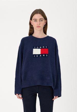 Suéter de punto azul marino con un cuello redondo, de corte oversized y puños acanalados. Presenta un logotipo central de "TOMMY JEANS" en colores contrastantes.