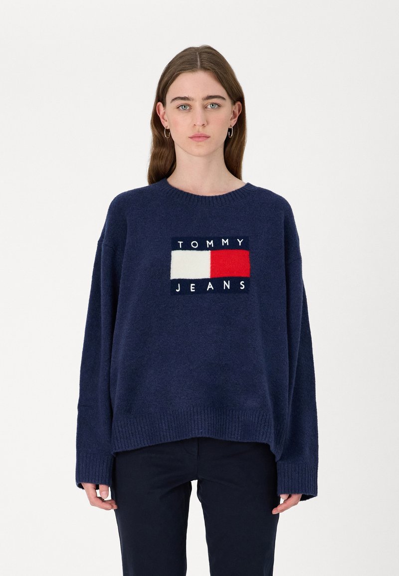 Maglione lavorato a maglia navy con scollatura rotonda, vestibilità oversized e polsini a coste. Presenta un logo centrale "TOMMY JEANS" in colori a contrasto.