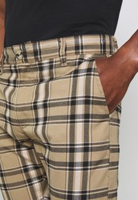 Beige plaid trousers with black and white accents, featuring belt loops, a front pocket, and textured fabric. Metallic button detail visible.