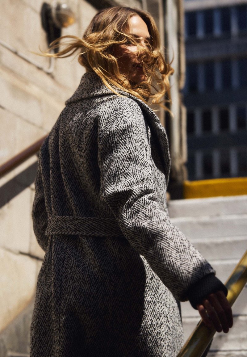 Grijze herringbone wol coat met een ceintuur in de taille, brede kraag en geribde polsmanchetten, gedragen terwijl hij de trap op loopt. Natuurlijk licht verlicht de scène.