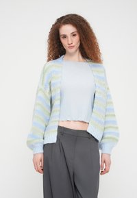 Πολύχρωμο πλεκτό ζακέτα με ριγέ σε παστέλ μπλε και πράσινο, σε υπερμεγέθη εφαρμογή και με rib μανσέτες. Στυλιζαρισμένο πάνω από ανοιχτό μπλε cropped τοπ και γκρι παντελόνια.