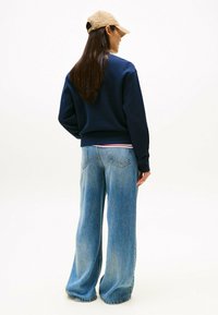 Felpa navy con polsini a costine, abbinata a jeans a gamba larga blu chiaro. Il modello indossa un cappellino beige, mostrando un look casual e rilassato.