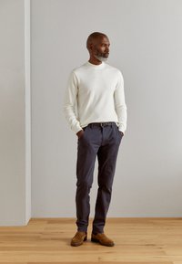 Pull long à manches blanches, pantalon bleu marine avec une ceinture, et bottes en daim marron ; pose debout contre un mur gris clair avec un sol en bois.