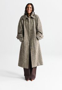 Manteau long en pied-de-poule marron avec un grand col, fermeture à boutons et manches retroussées. Porté sur un pantalon marron foncé et des chaussures pointues.