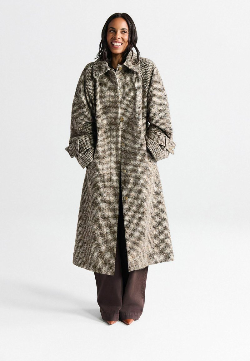 Manteau long en pied-de-poule marron avec un grand col, fermeture à boutons et manches retroussées. Porté sur un pantalon marron foncé et des chaussures pointues.