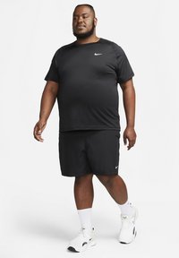 Camiseta y pantalones cortos negros de Nike que absorben la humedad. Logo de Nike en blanco en la camiseta y en las zapatillas. El modelo lleva calcetines blancos con zapatillas deportivas de suela blanca.