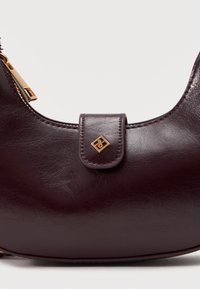 Σκούρο μοβ δερμάτινη τσάντα χειρός με ημισέληνο σχήμα, χρυστό φερμουάρ και μικρό χρυσό τετράγωνο έμβλημα σε μπροστινή στερέωση ιμάντα.