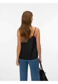 Svart satengcamisole med tynne stropper, avslappet passform, kombinert med lyseblå vidbukser. Modellen holder en svart handlenett i høyre hånd.