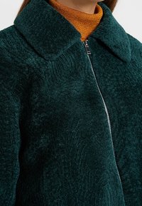 Tearsfarvet tekstureret jakke med lynlås, stor krave og et mønster der minder om bølger. Under er en gylden turtleneck synlig.