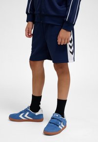 Gyermek sötétkék rövidnadrágban, fehér oldalsávos, hozzáillő kabátban, fekete zokniban és kék tépőzáras sportcipőben, fehér részletekkel.