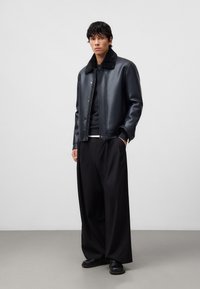 Uomo che indossa una giacca nera in ecopelle con colletto di pelliccia, pantaloni neri a gamba larga, maglione nero e scarpe nere, in piedi contro uno sfondo grigio.